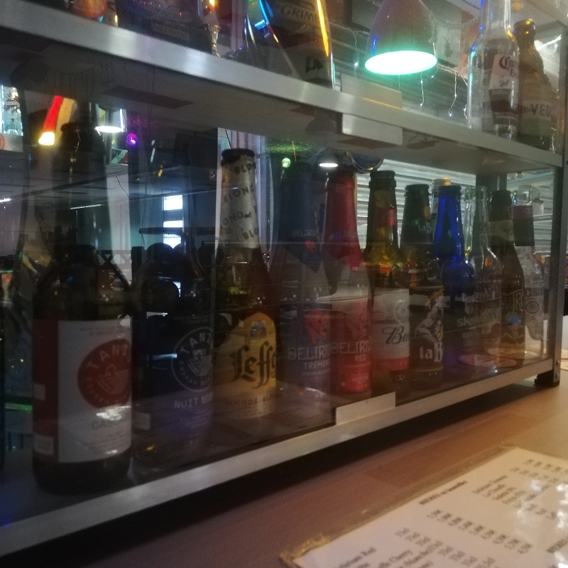 Vitrine en verre remplie de bouteilles de bière variées, dont Leffe Blonde, Delirium Tremens, Budweiser. Un menu est visible en bas.