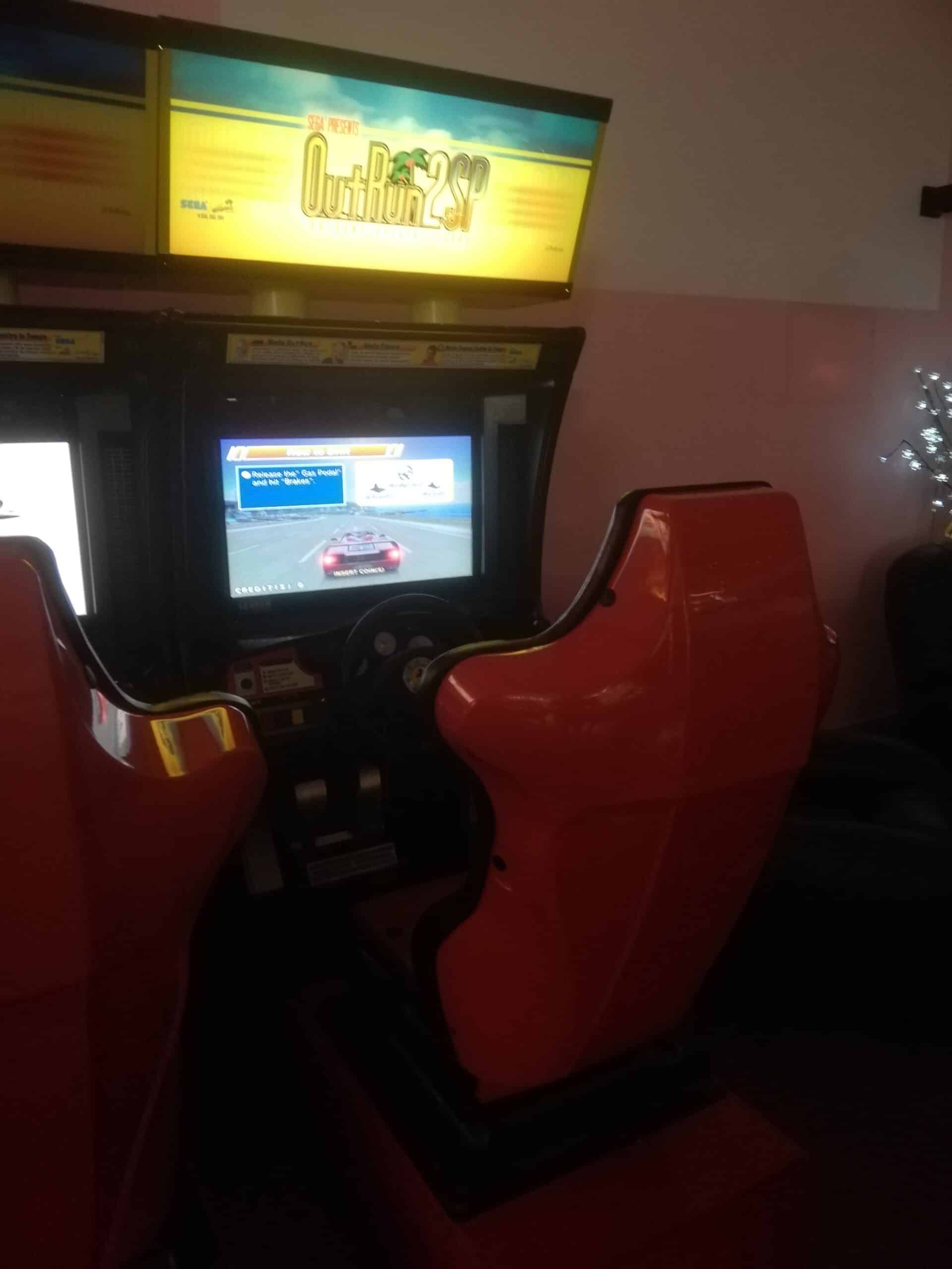 Borne d'arcade Sega OutRun 2 SP avec deux sièges rouges, l'écran affiche une voiture de course et les instructions 'Release the Gas and hit Brakes' et 'INSERT COIN(S)'.