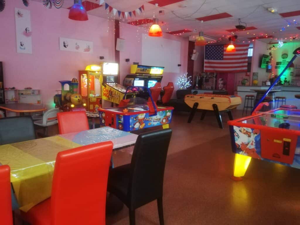 Salle de jeux d'arcade festive: tables, chaises rouges, baby-foot, air hockey, train enfant et décorations américaines lumineuses.