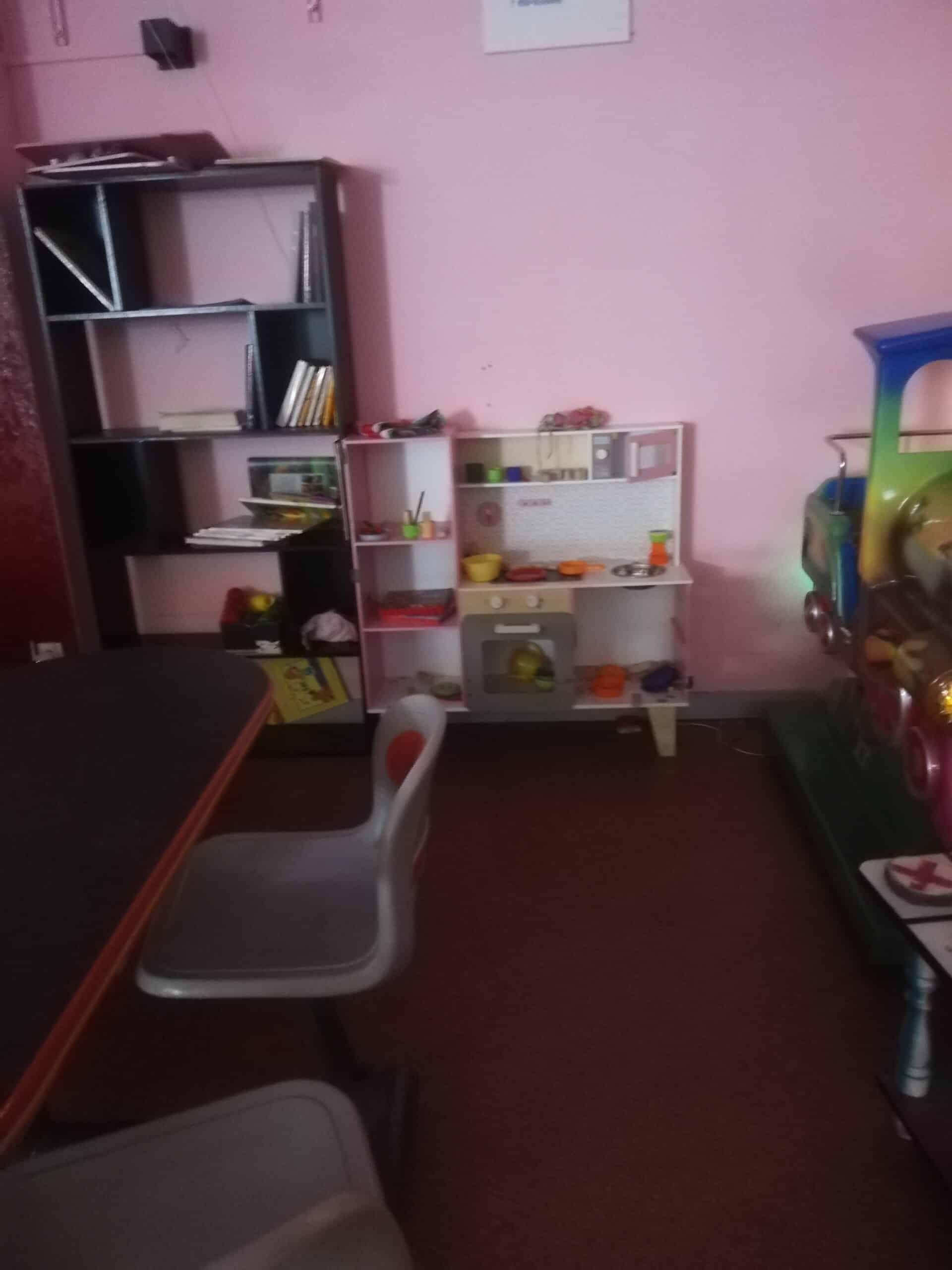 Salle de jeu enfantine aux murs roses. Une étagère de livres côtoie une cuisine jouet et un train coloré. Table et chaise visibles.